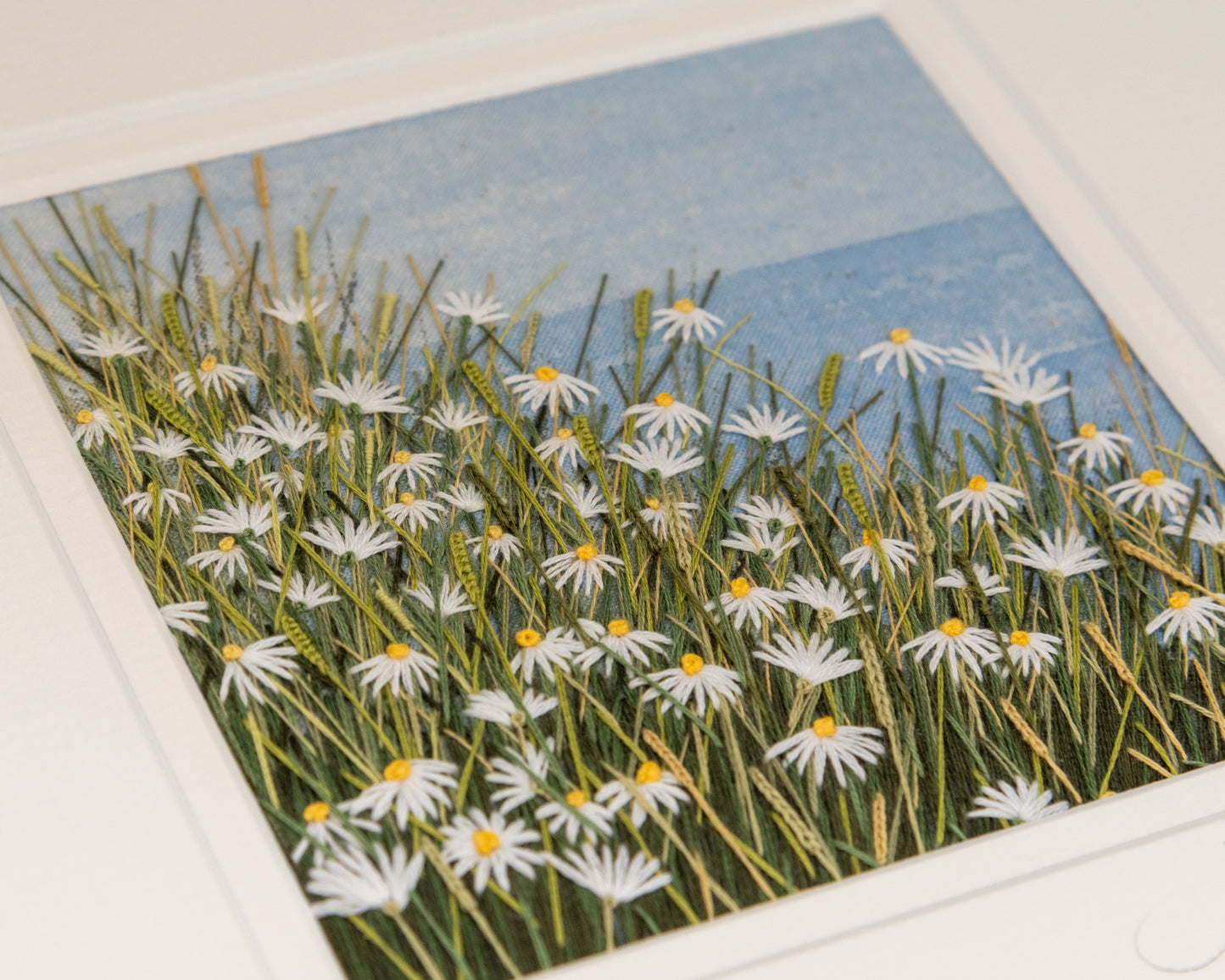 Framed embroidery artwork of a field of daisies with a painted horizon and sky background