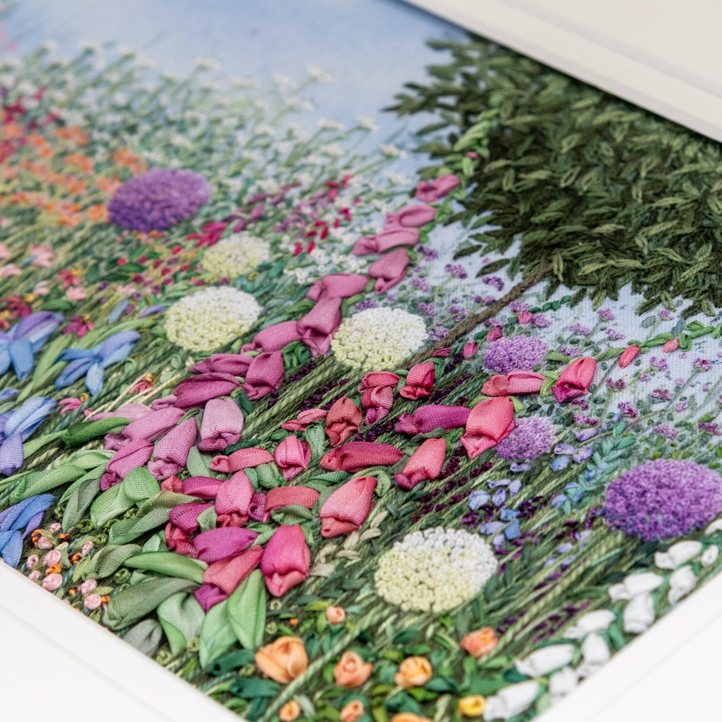 Close up of a hand embroidered garden brder. The main feature is the bottom half of a round bay topiary tree underneath sits a lush flower border of alliums, foxgloves, irises, verbena bonairensis, roses and other flowers and foliage.