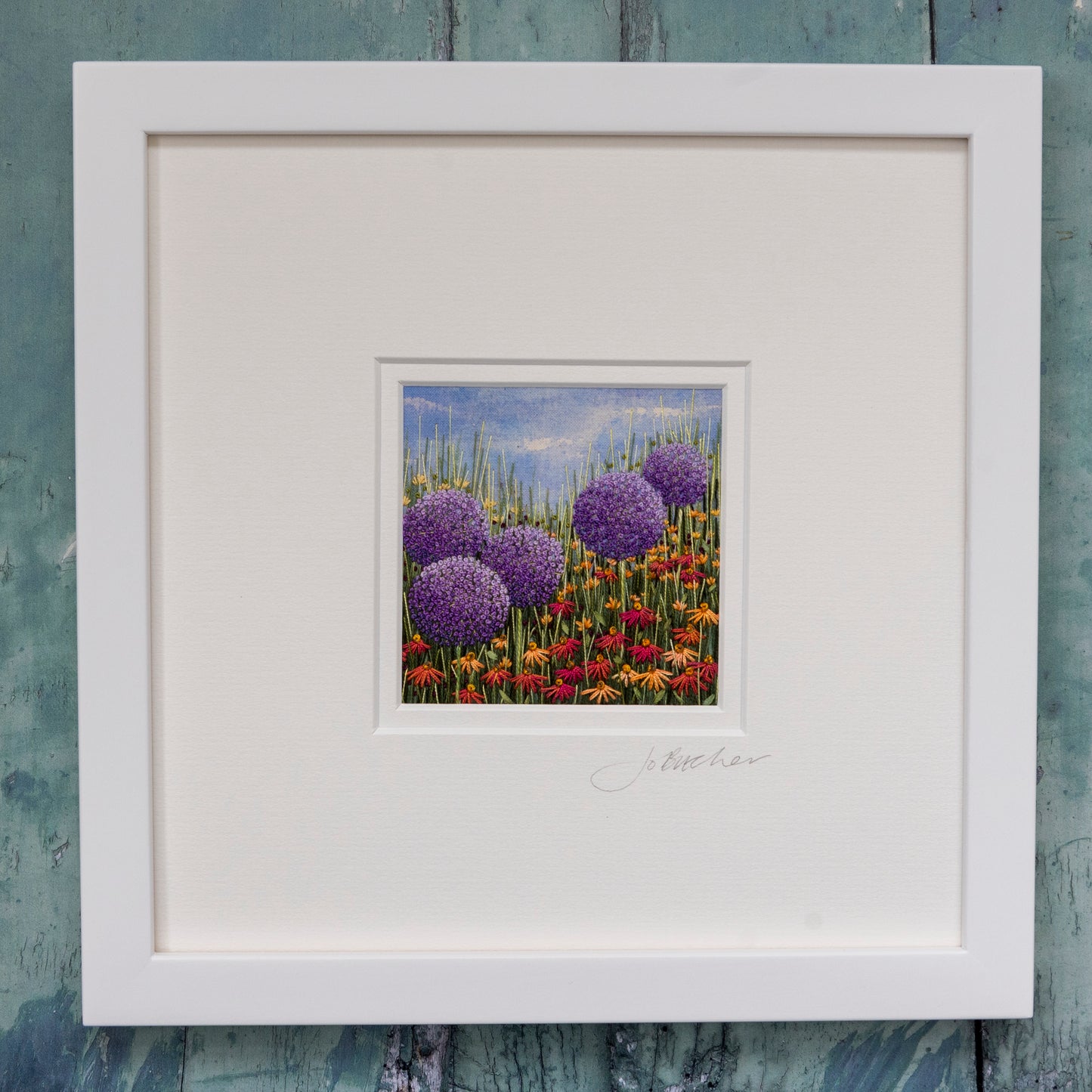 This framed hand embroidered artwork by Jo Butcher is of five large purple allium heads.
The allium heads are sitting above red/orange daisies, grasses and foliage