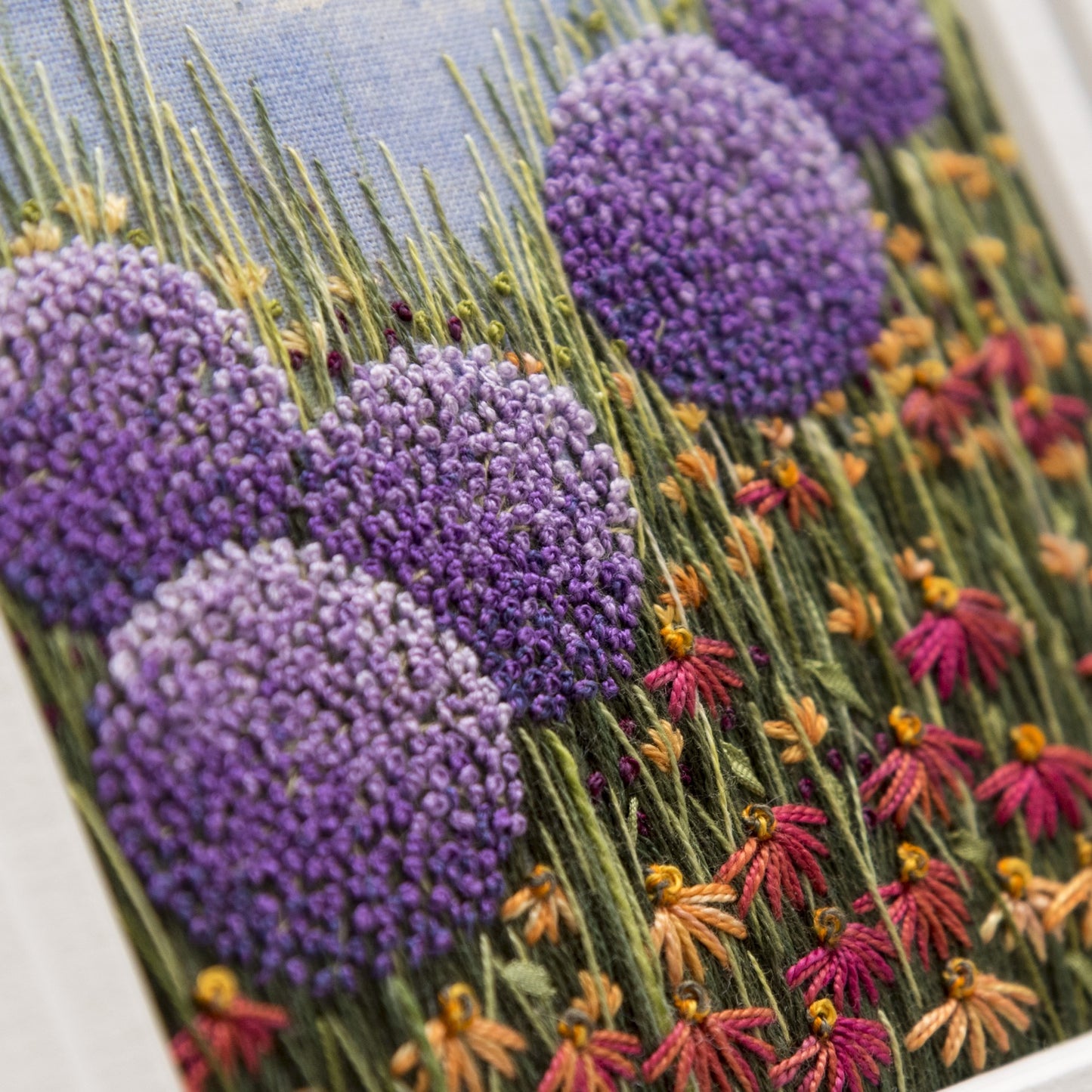 Five large purple allium heads are the main feature of this hand embroiery artwork by Jo Butcher.  
The allium heads are sitting above red/orange daisies, grasses and foliage