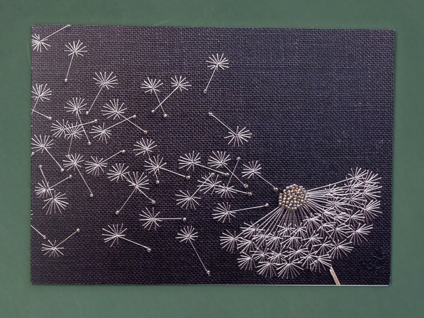 A charcoal grey background with white dandelion clock hand embroidered with the effect of the wind blowing the seeds away.