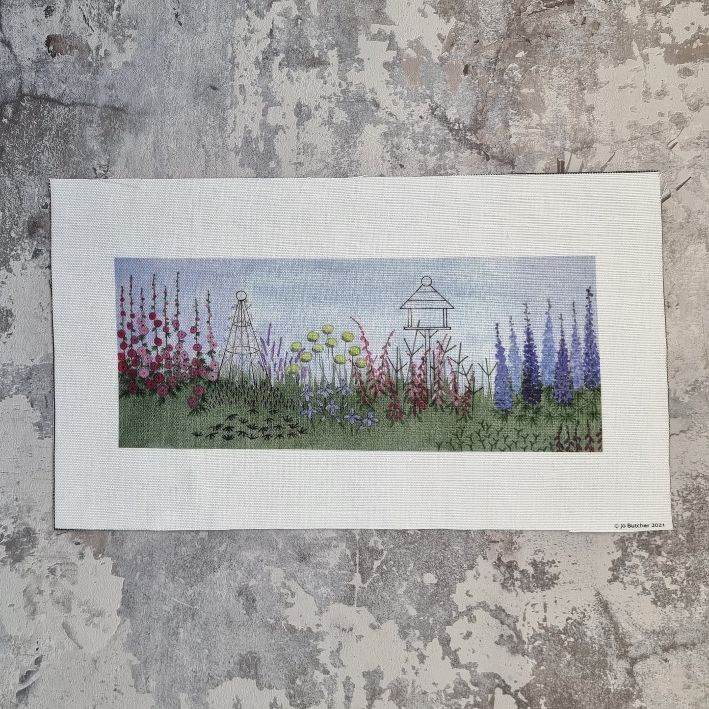 A rectangular piece of printed cotton linen.  The printed design is of a herbaceous border with a bird table and rose obelisk rising above the planting of foxgloves, delphiniums, hollyhocks and other plants and foliage.