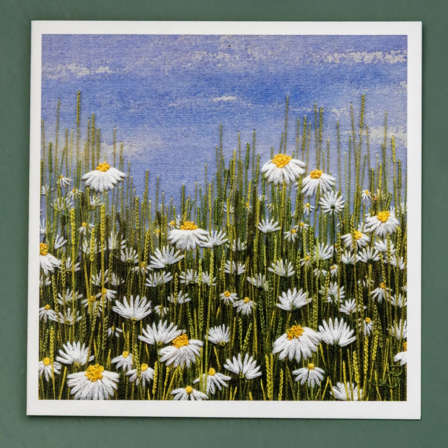 a card of a hand embroidered daisy meadow on a painted sky background.