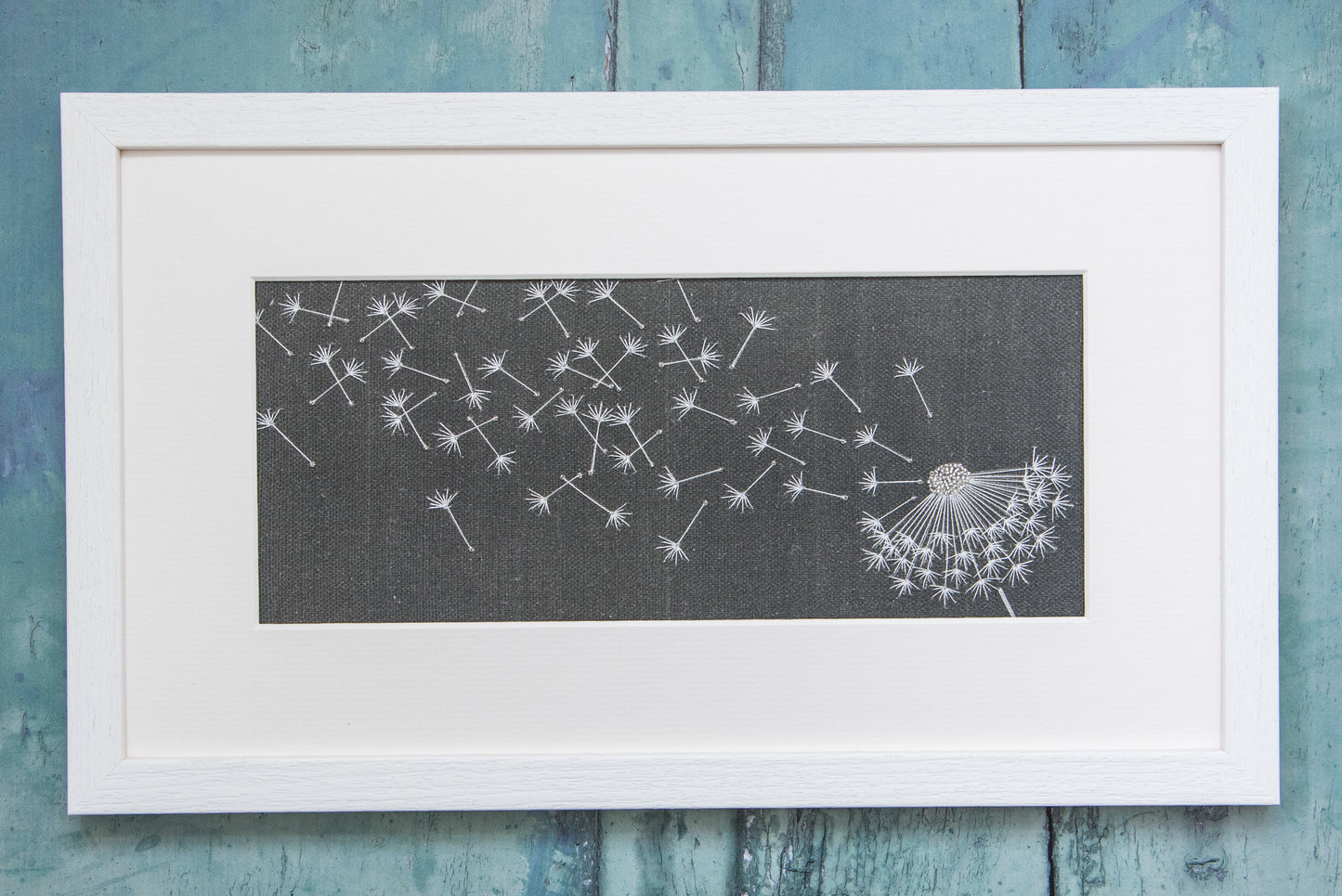 Framed finished embroidery of a dandelion clock caught in the wind on a charcoal cotton/linen background.