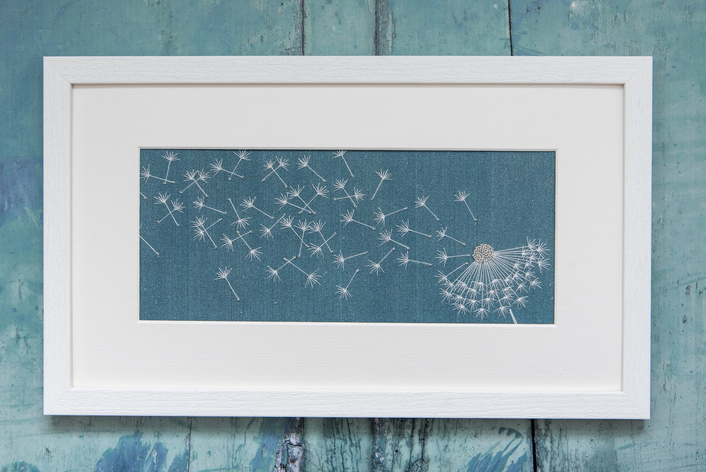 A petrol blue background with a embroidered dandelion clock blown by the wind. Framed in a white frame.