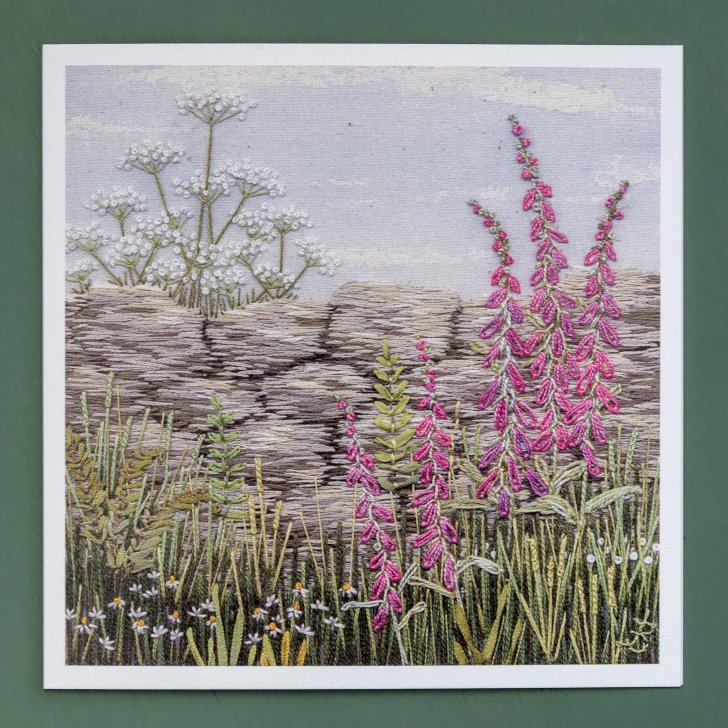 A hand embroidered classic scene of a walk in the country. The main feature is a  dry stone wall with pink foxgloves in front of the wall and  cow parsley rising behind. The foreground is grasses, daises and ferns.