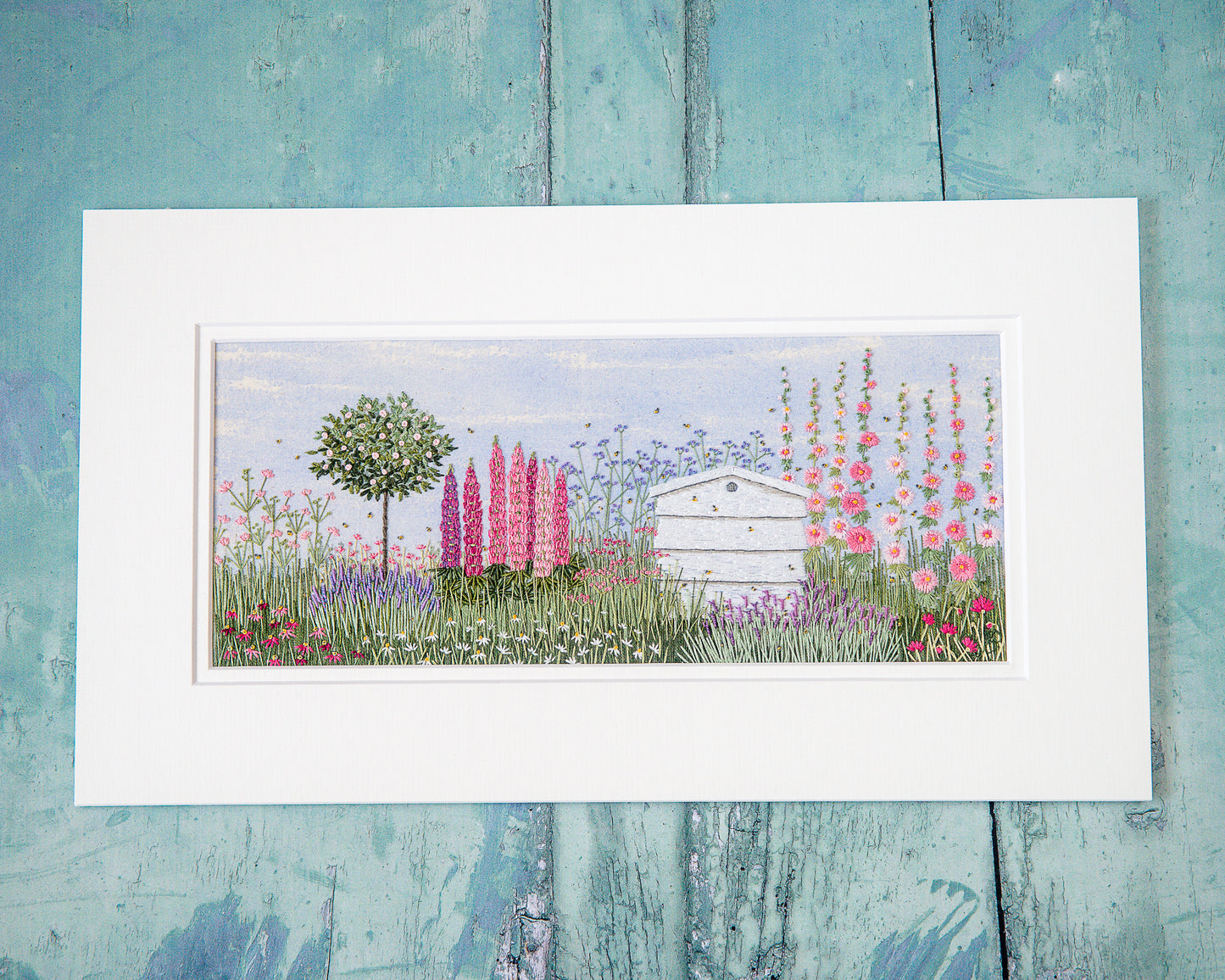 a white wooden beehive sits amongst a flower border full of colour.  Bees are buzzing around in this complete hand embroidery kit.