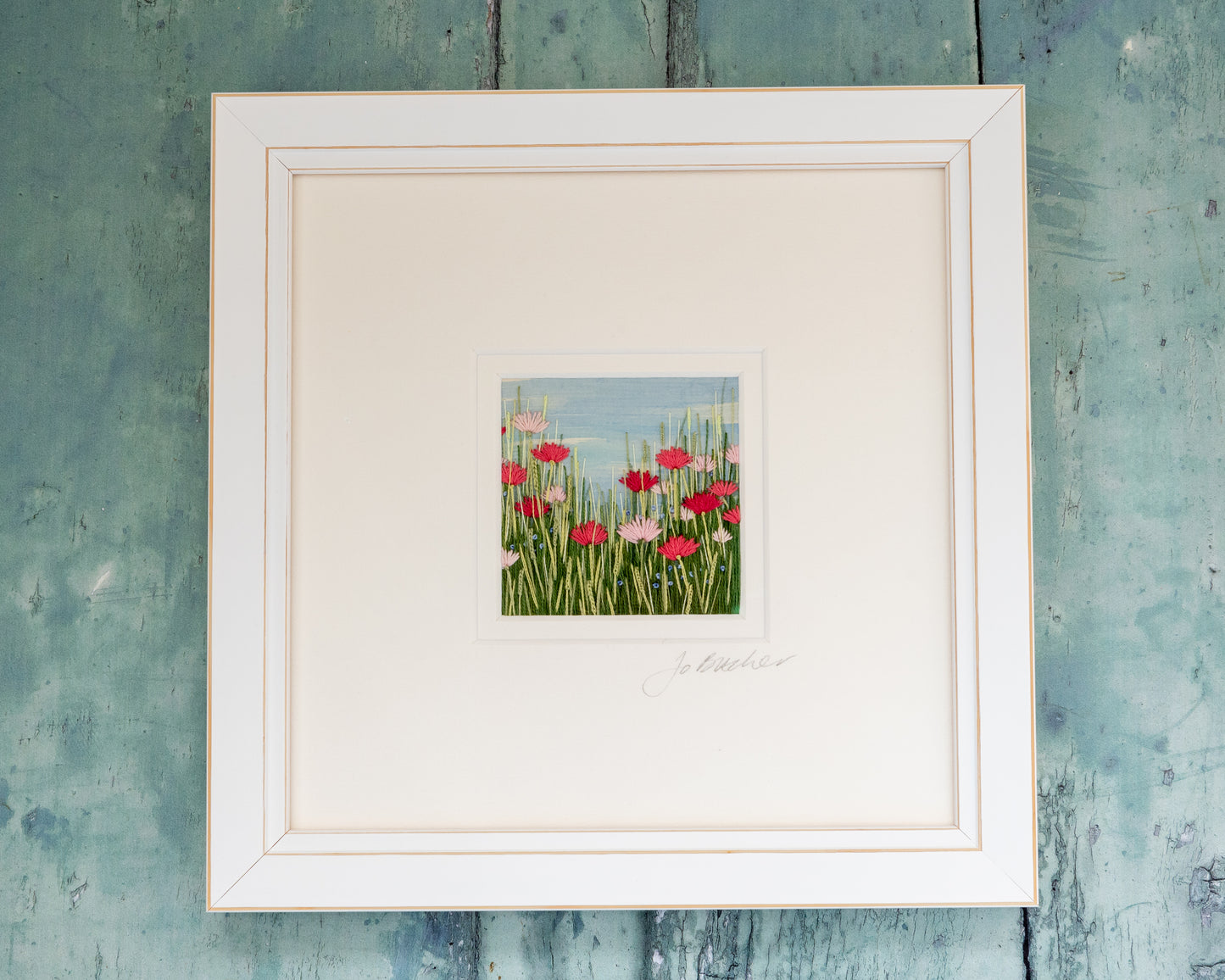 White framed hand embroidered artwork featuring poppy like flowers in shades of pink and small blue flowers amongst he grasses.