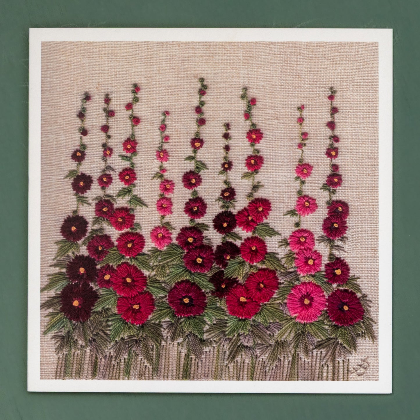 card showing nine hand embroidered hollyhocks. The hollyhocks range in colour from deep purple to bright pink.