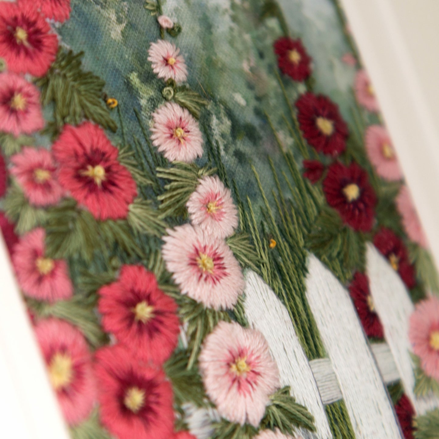 A white embroidered picket fence is seen surrounded by spires of hollyhocks in shades of pink.