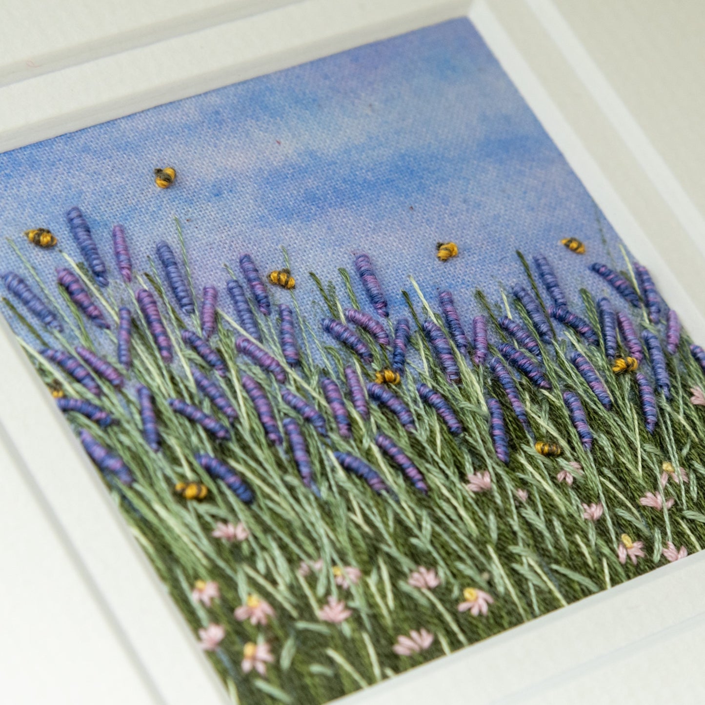 Hand embroidered artwork of bees buzzing around the lavender with grass and pale pink daisies in the foreground