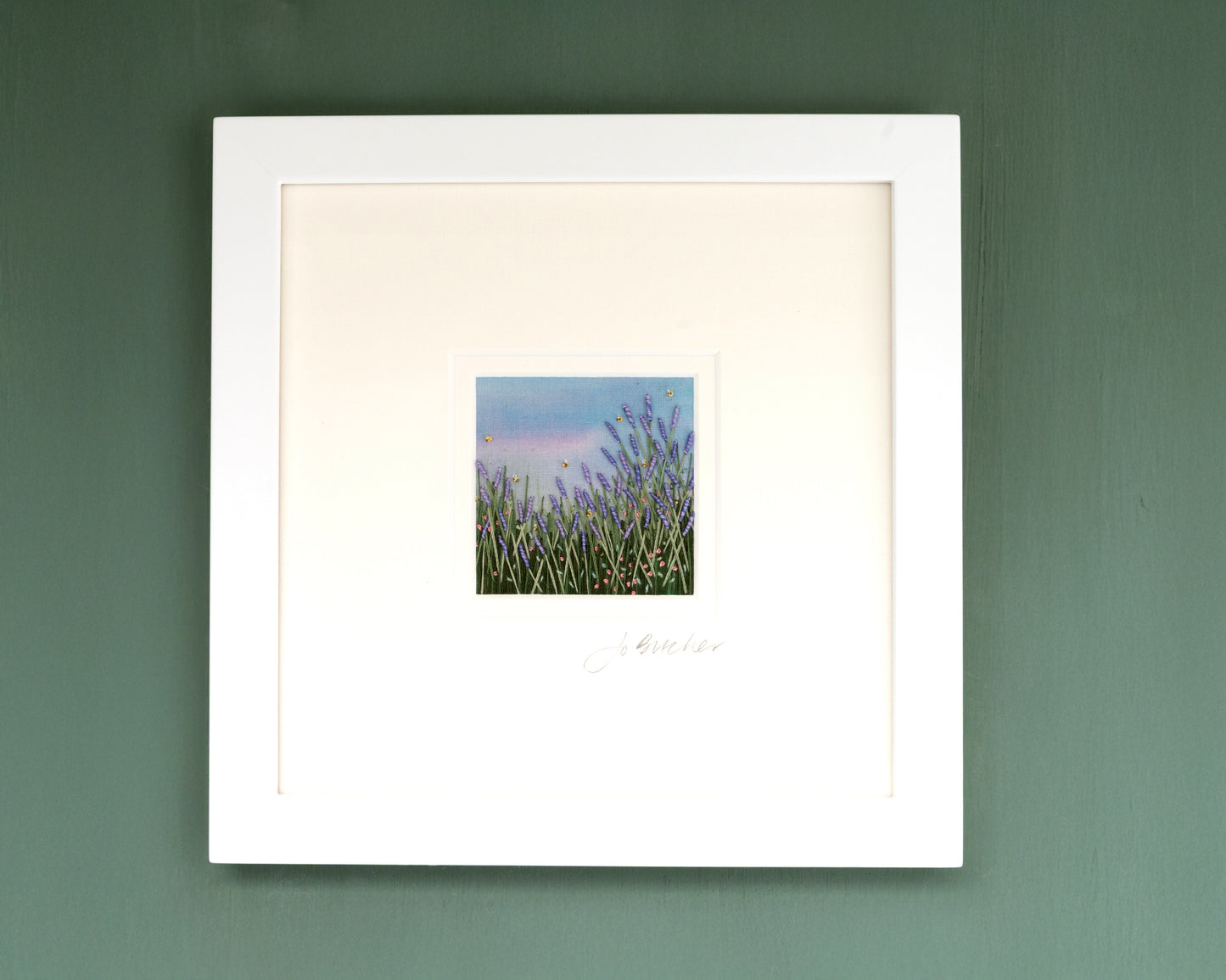 A white framed hand embroidery artwork of Lavender and pink flowers amongst the grass and foliage. A few bees are also included.