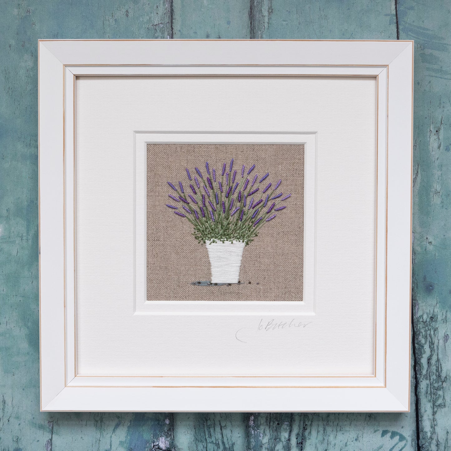 A white framed artwork of a flowering  Lavender plant growing in a white pot on a beige cotton/linen background.