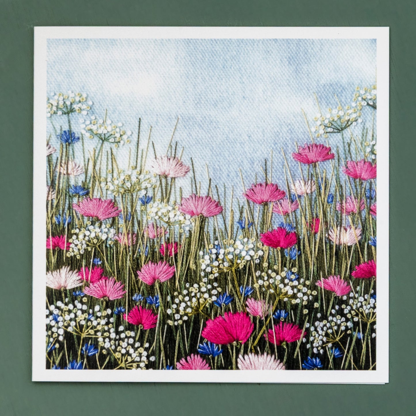 A card of a hand embroidered grass meadow from a rabbits eye view.  Among the grasses are pretty blue cornflowers and pink poppies.