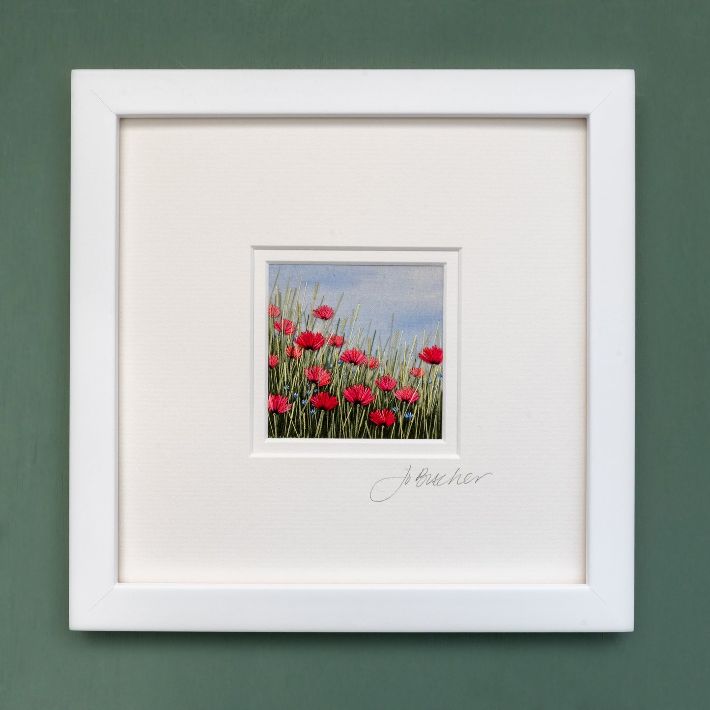 A white framed artwork of a red/pink poppy meadow.  A few small blue flowers and amongst he flowering grasses.