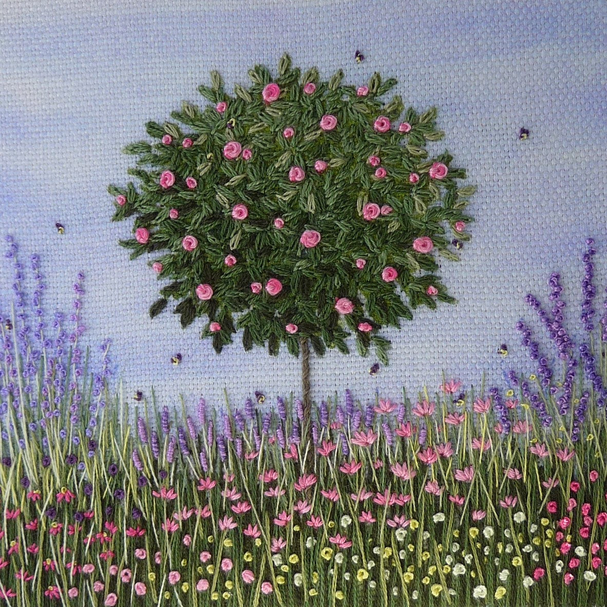 A standard rose tree stands above a flower border with pink roses in bloom and bees busy flying around the flowers