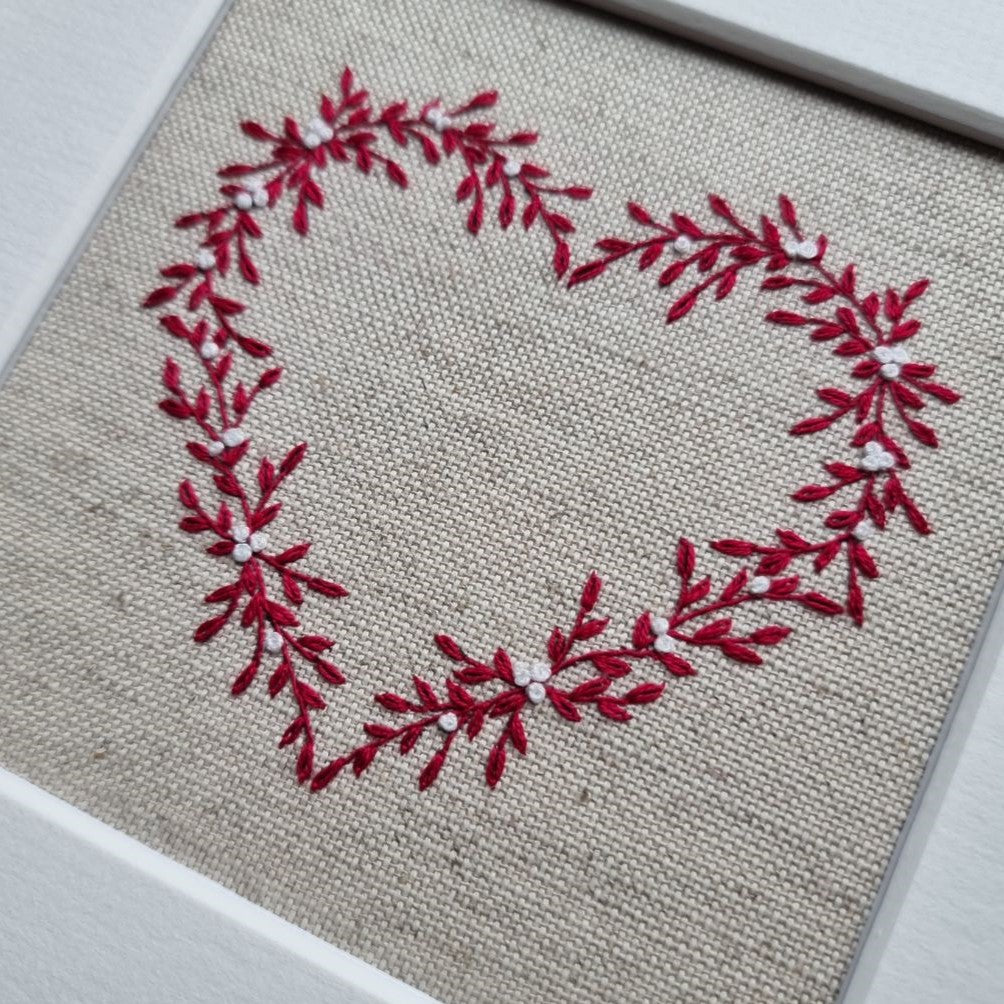 This embroidery kit is a heart stitched of red leaves and white berries on a beige linen background