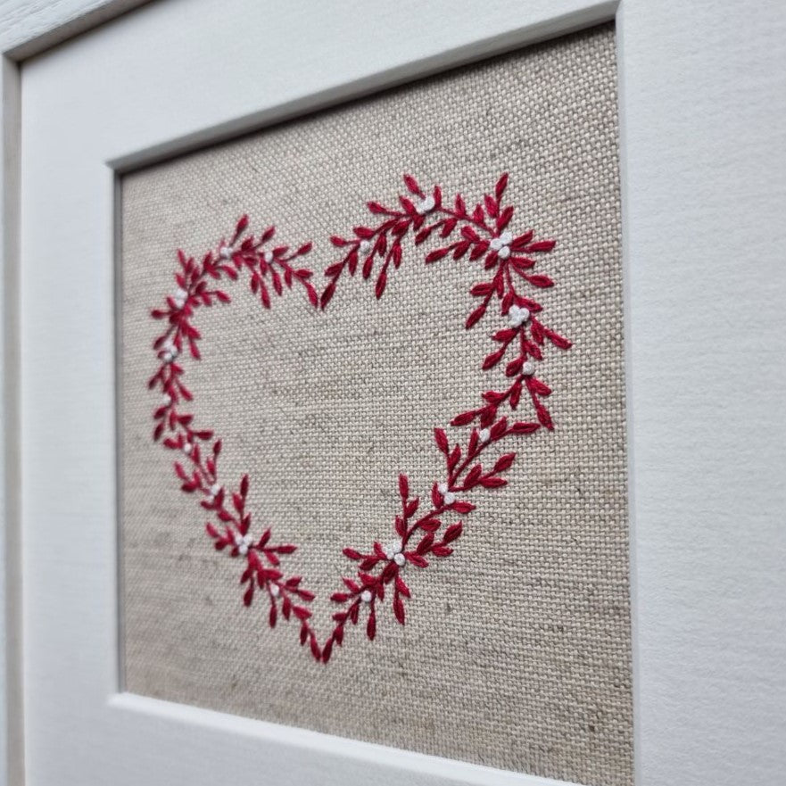 This embroidery kit is a heart stitched of red leaves and white berries on a beige linen background