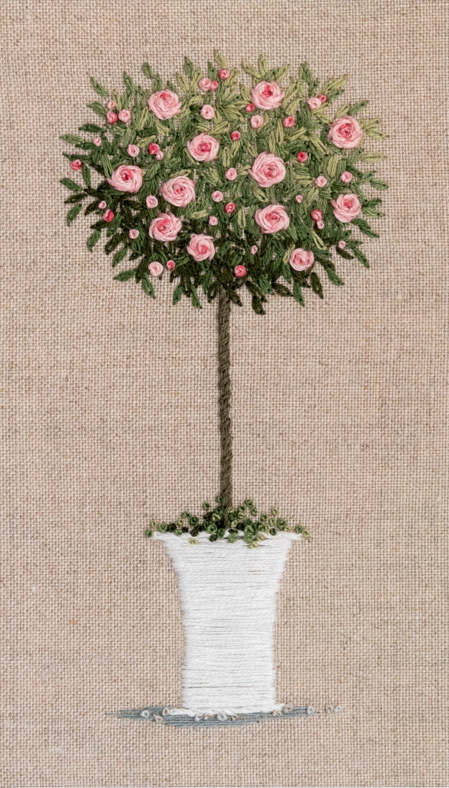 Hand embroidered white pot has rose tree  with pink flowering roses and buds. The background is beige cotton/linen.