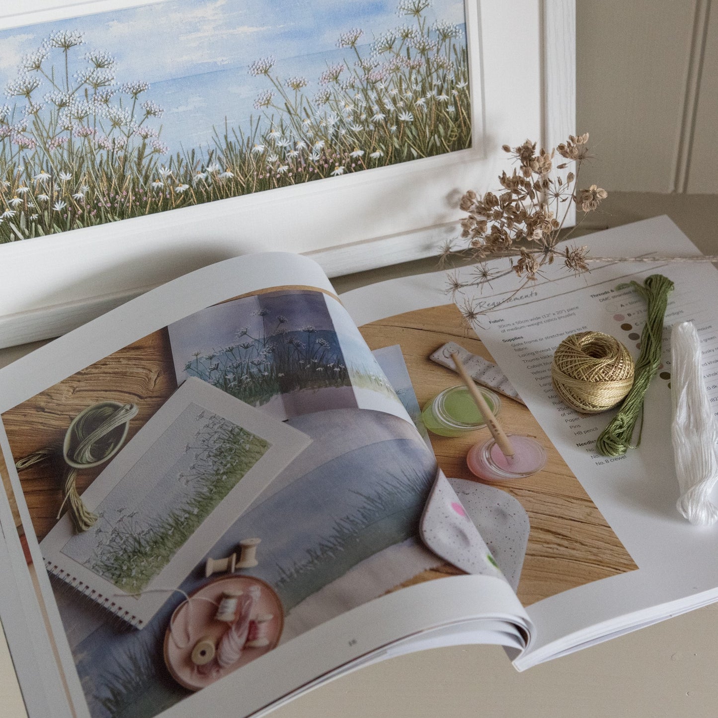 A book is open at a page showing the images of the seascape project with a few skeins and balls of cotton and a dried caw parsley head.  The original artwork of a seascape depicting the grasses and daises on a coastal path is propped up in the background