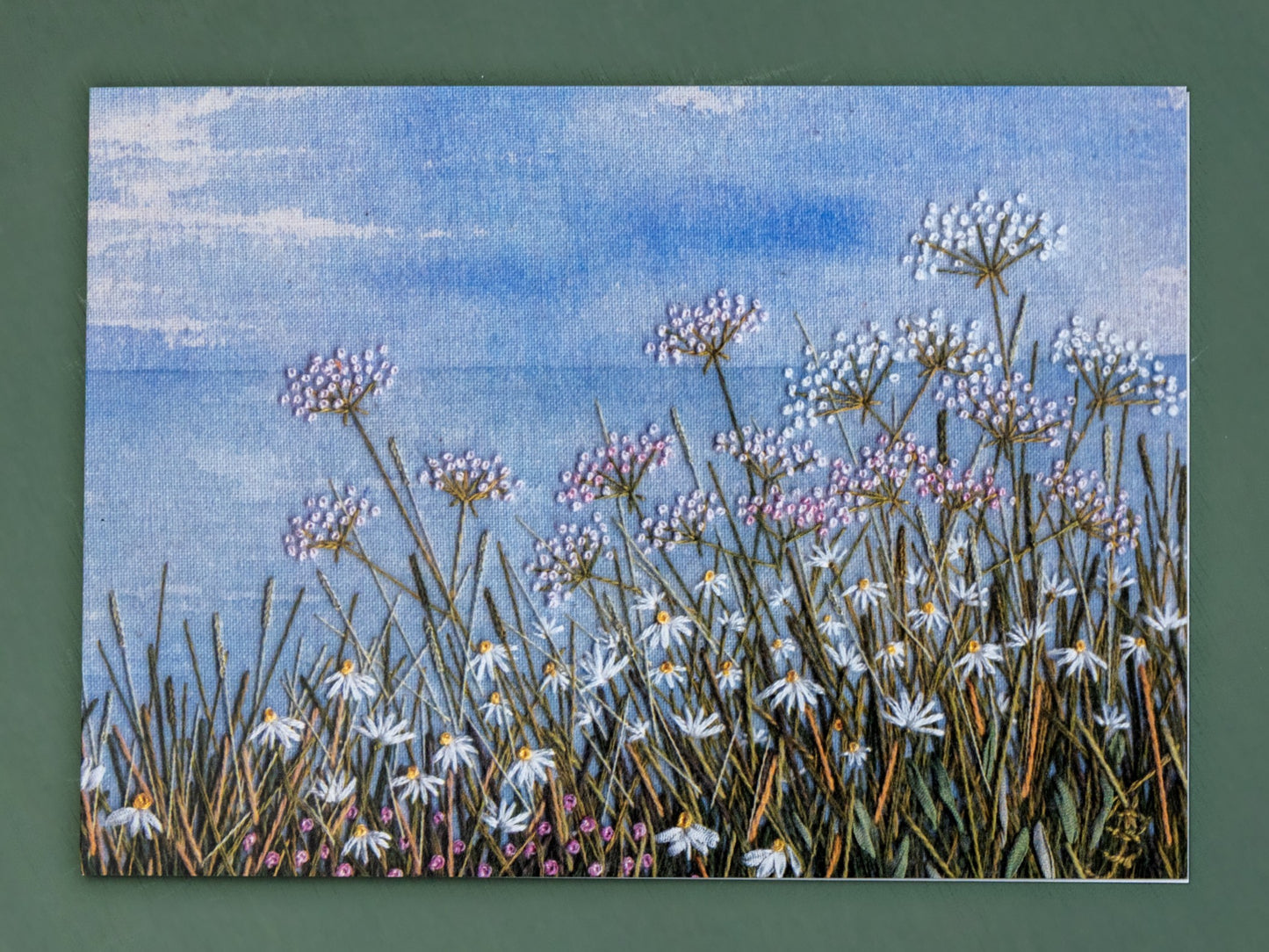 A card of a classic coastal scene. The hand painted  background shows the horizon and the foreground is coastal wild flowers.