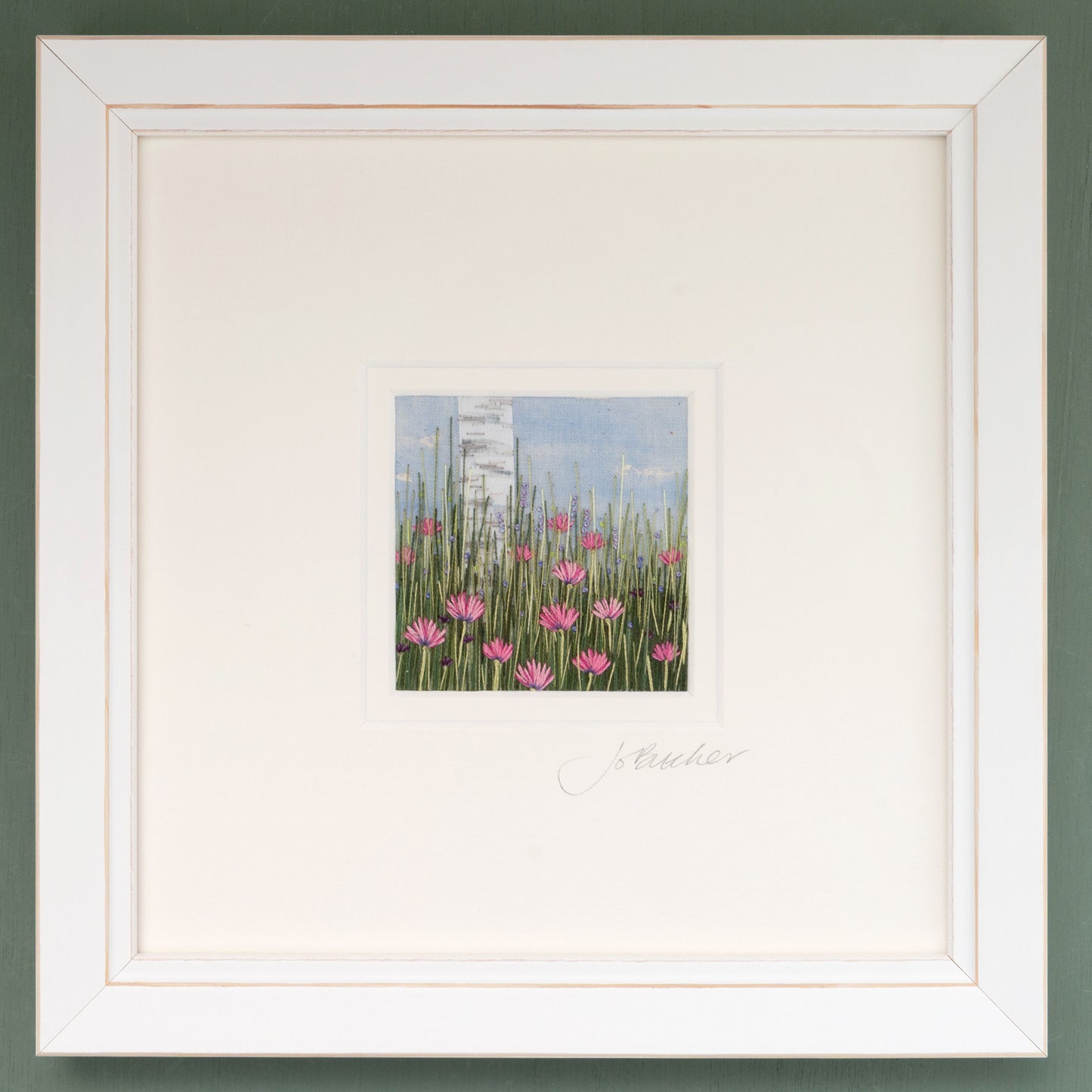 A white framed embroidery of a silver birch trunk rising above pretty pink flowers. Smaller purple and blue flowers nestle in the grasses.
