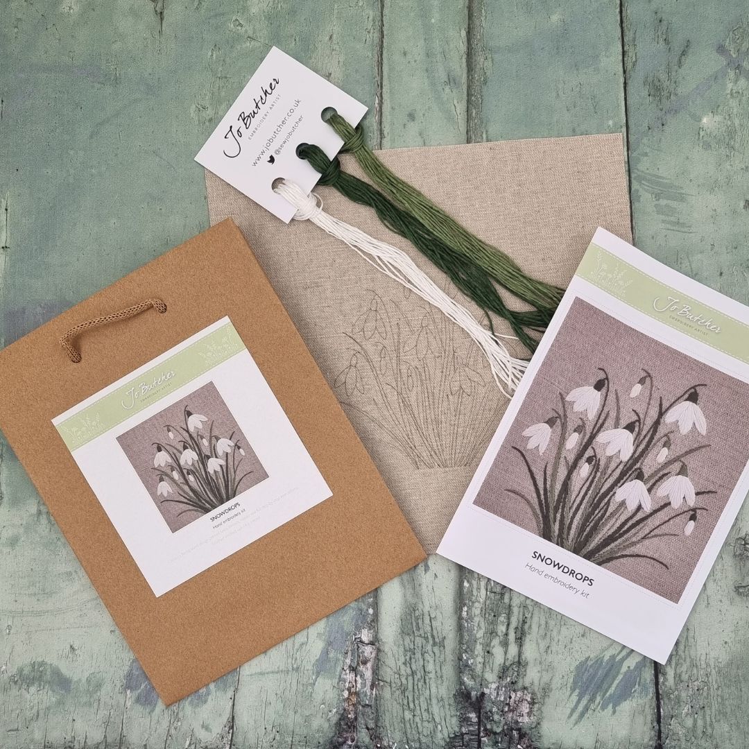 the contents of the snowdrops kit by Jo Butcher are shown to be a printed linen background, threads on  thread card, the image of the front of the instructions and the brown paper bag the kit comes in.