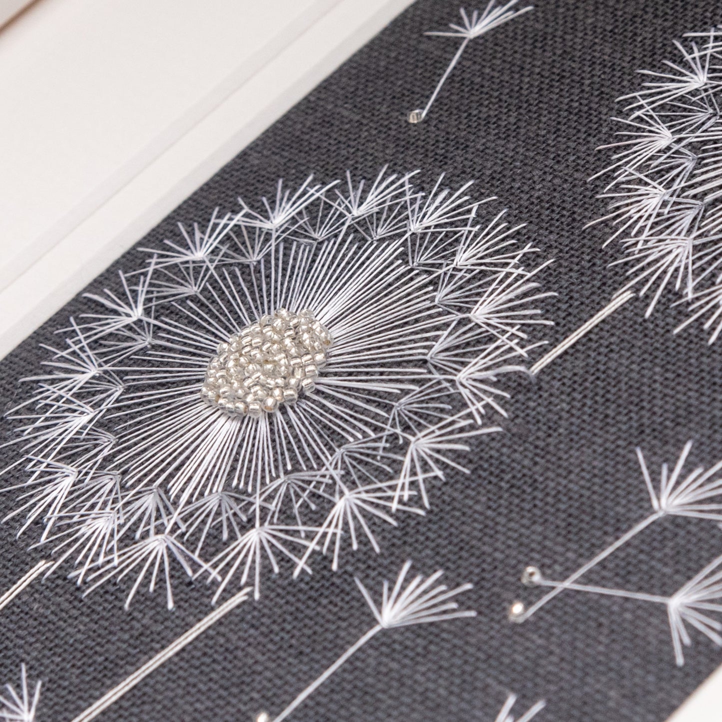 embroidered close up of dandelion seed head on a charcoal background using glass beads and white thread