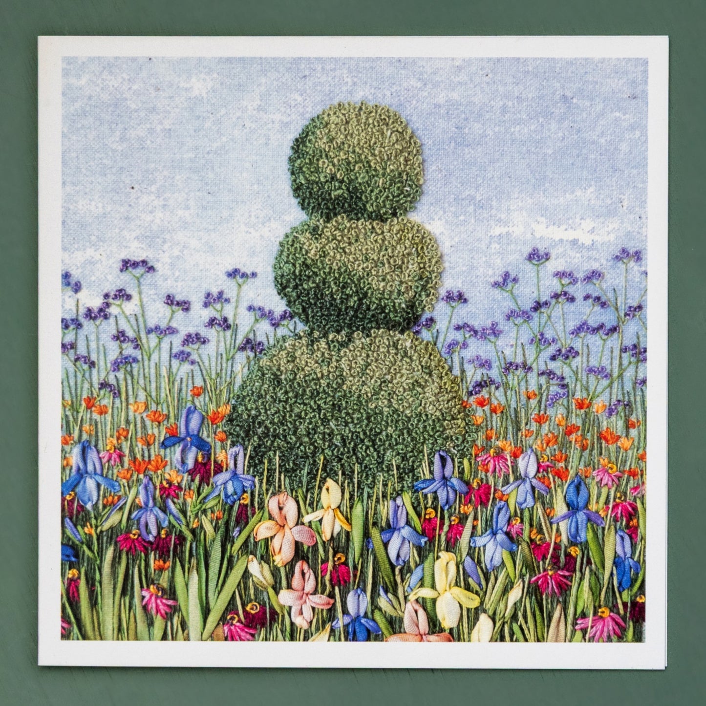 A hand embroidered three ball topiary tree rises above a flower border of a ribbon embroidered irises and hot orange flowers with tall verbena bonariensis in the background.
