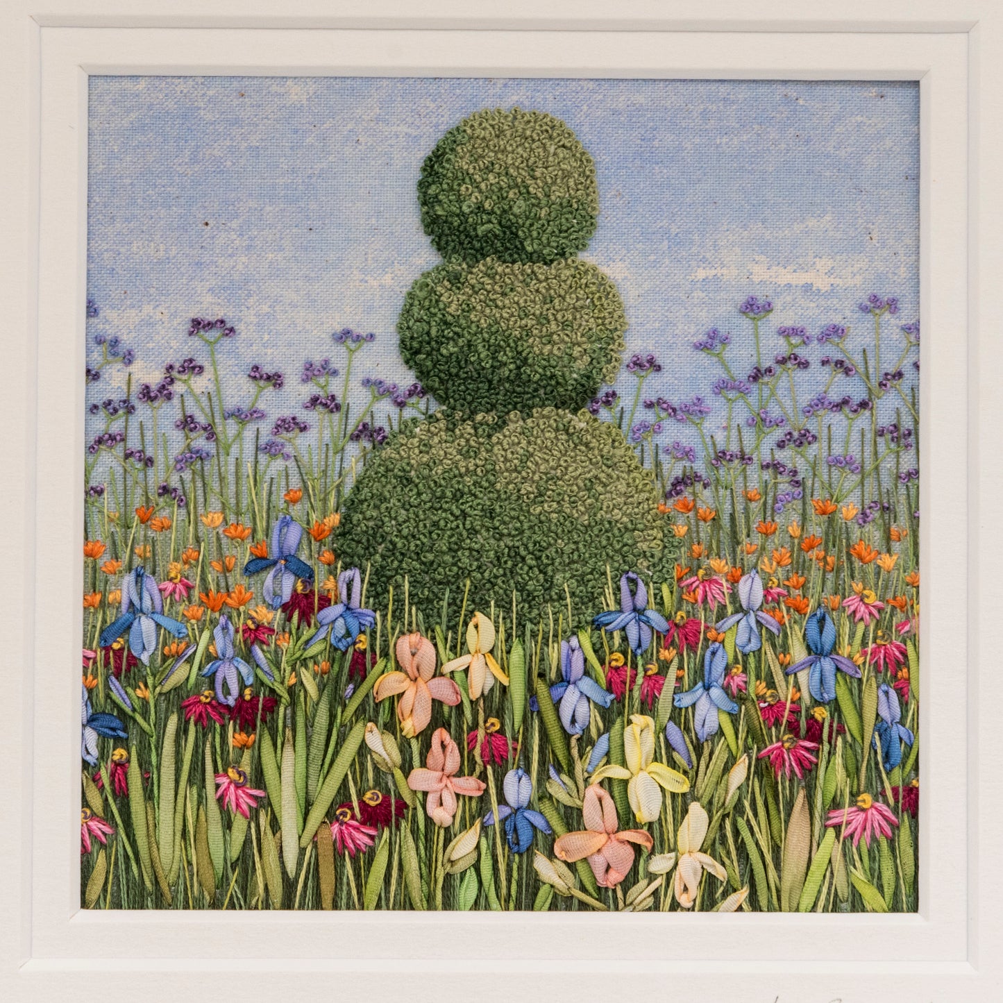 hand embroidery of a garden border with a topiary feature of three decreasing size balls on top of each other. The foreground shows Irises and foliage and verbena bonairensis gives some background height