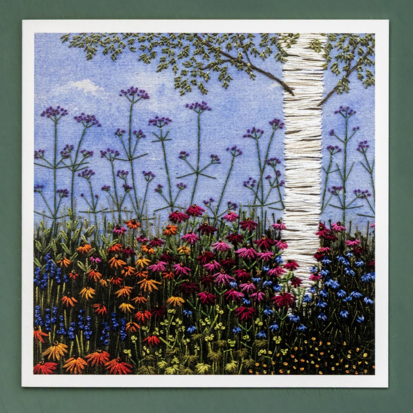 A hand embroidered silver birch tree with lower branches is set in flowering border of deep purple and range echinacea and delicate blue and yellow flowers to contrast.