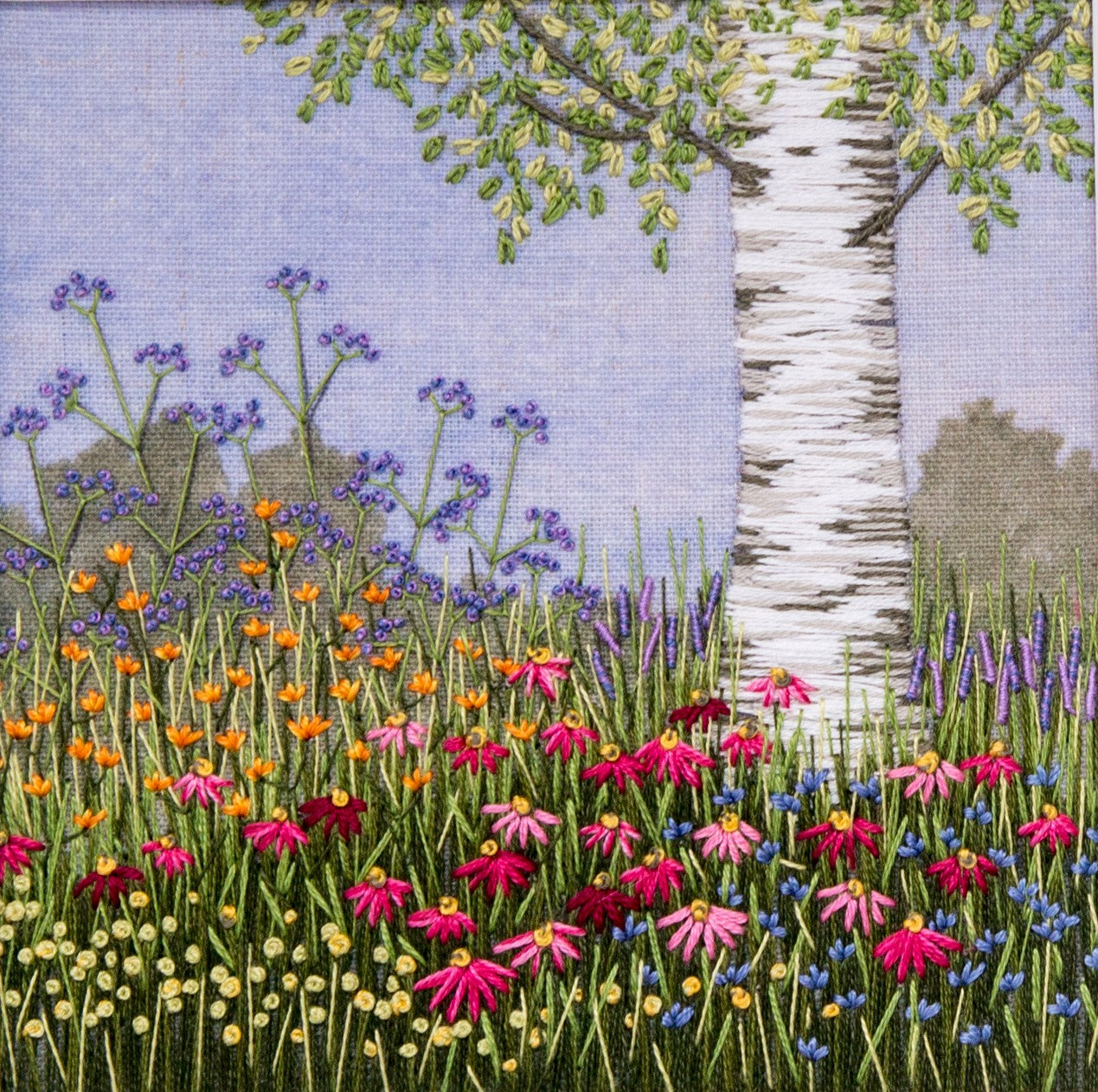 The image is of a silver birch trunk with the bottom of a few branches rising out of a mass of flowers of different colours. The background is printed with blue sky and a few hedges in the distance. it is a representation of what the the Under the silver birch tree embroidery kit should look like once completed.