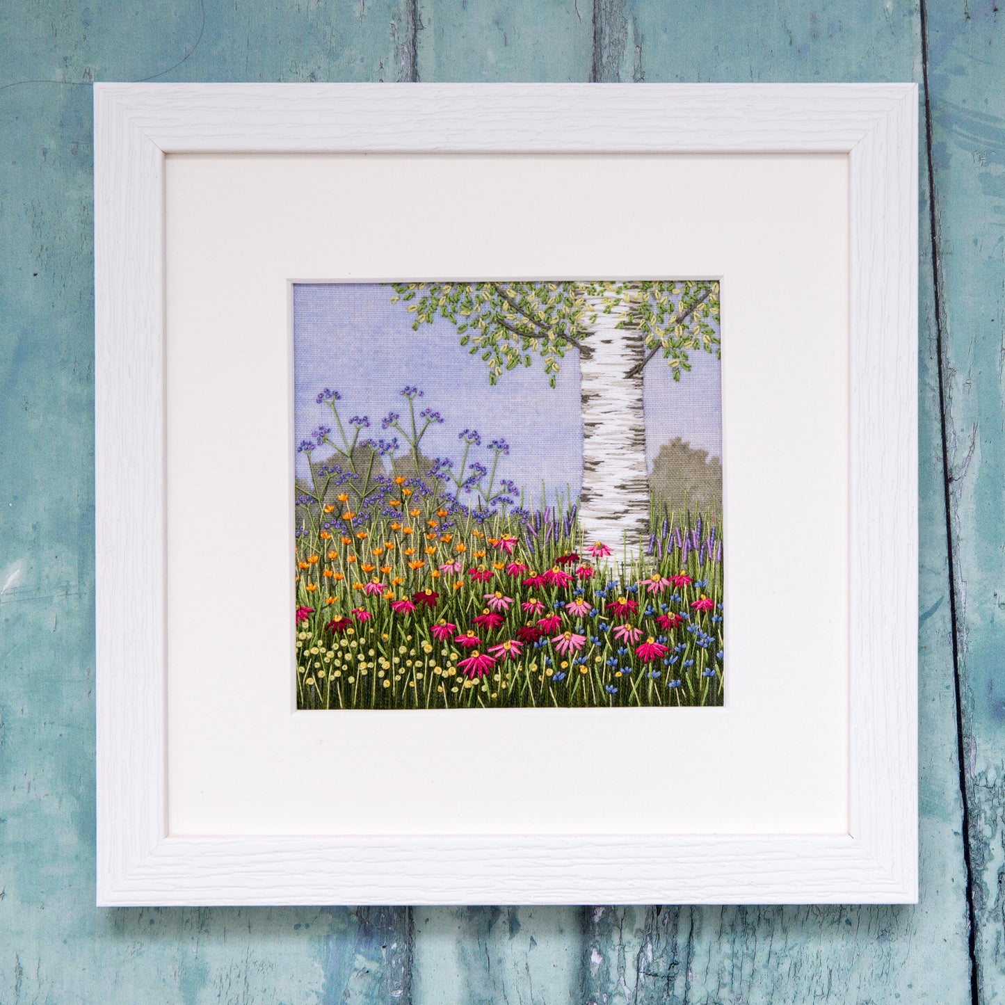 white framed artwork of a completed under the silver birch tree kit by Jo Butcher.  The image is of a silver birch trunk with the bottom of a few branches rising out of a mass of flowers of different colours. The background is printed with blue sky and a few hedges in the distance