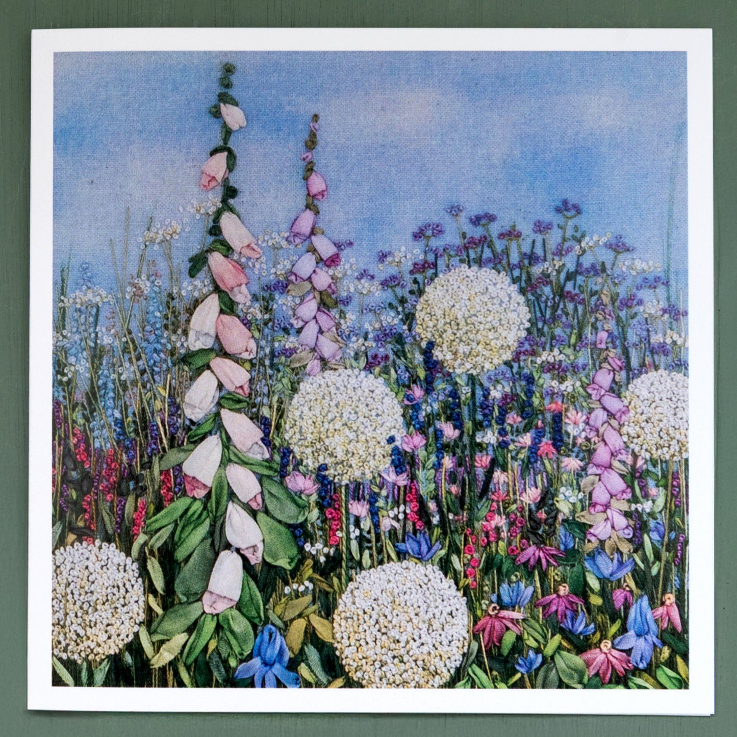 A square greeting card printed from an original Jo Butcher embroidery showing  five large white allium heads and pale foxgloves in a lush flower border.