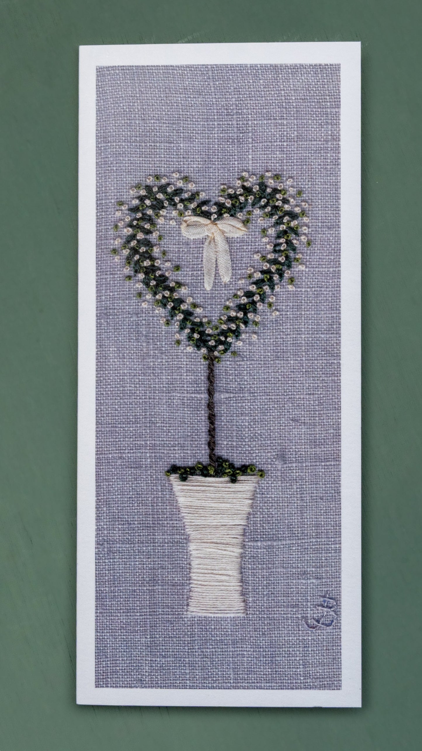 a hand embroidered white pot with a heart shaped tree with white roses and a ribbon bow hanging in the centre.