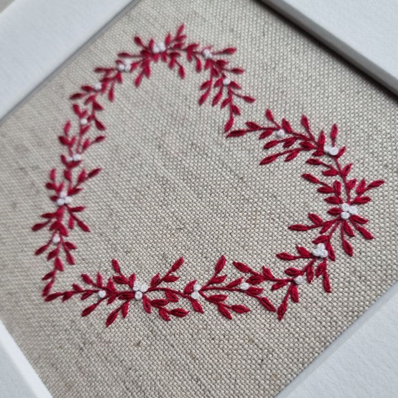 This embroidery kit is a heart stitched of red leaves and white berries on a beige linen background