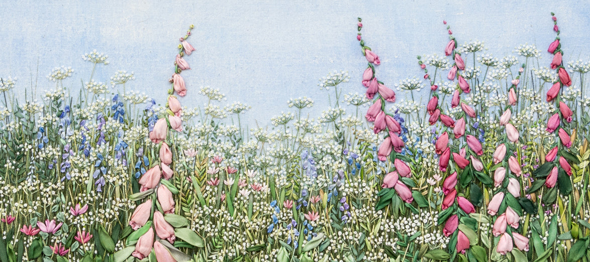 an intricately stitched flower border showing pink foxglove spires, blue hair bells and asters using silk ribbon amongst a sea of foliage and fine white flowers