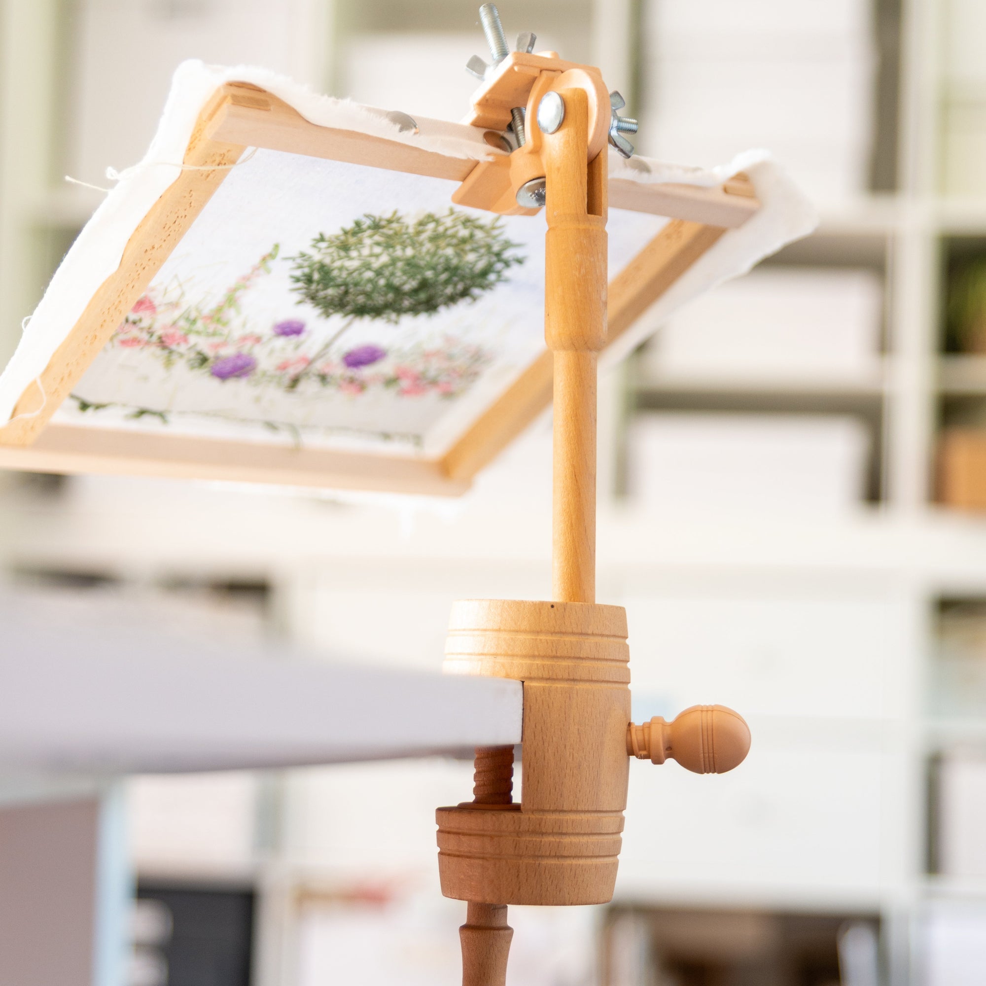 Wooden embroidery frame held in a table clamp with a small embroidery project on a blurred indoor background