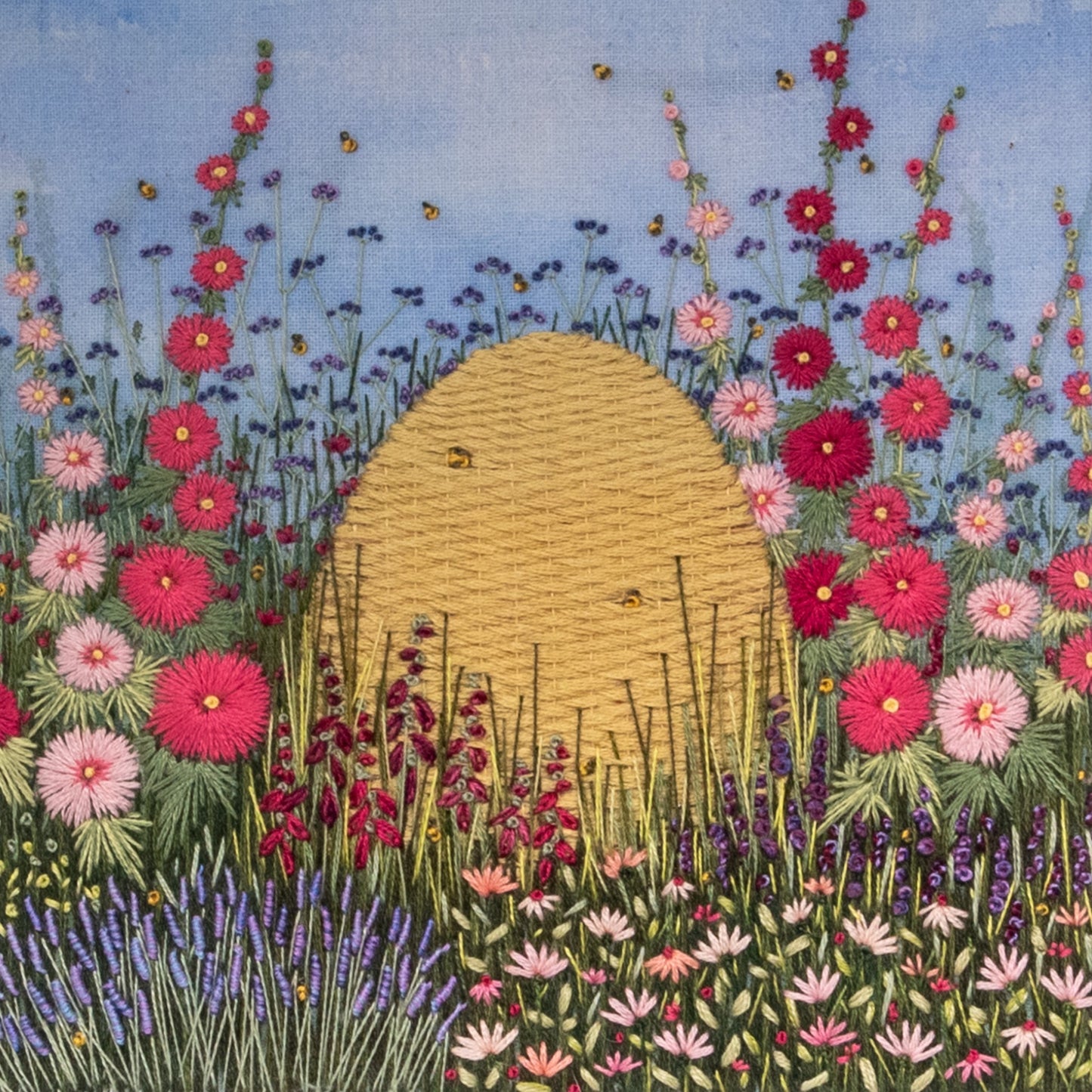 A intricate hand embroidered bee skep sit nestled with a lush flowering border of lavender, hollyhocks, salvias, foxgloves and cone flowers. A few bees buzz around 