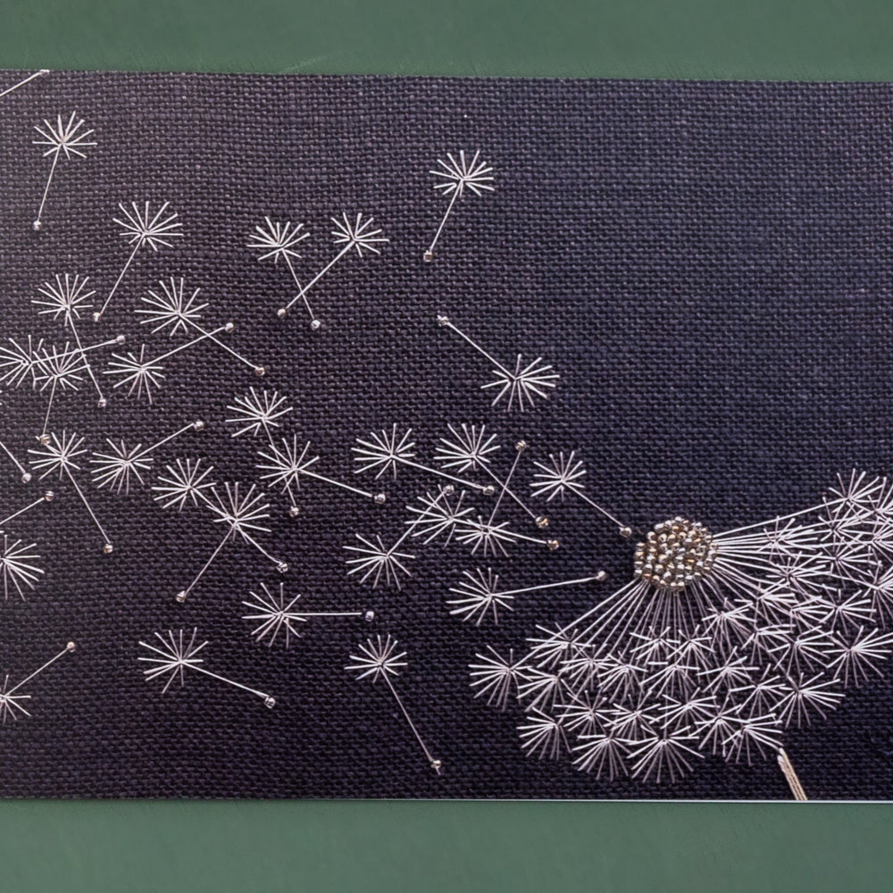 A charcoal grey background with white dandelion clock hand embroidered with the effect of the wind blowing the seeds away.