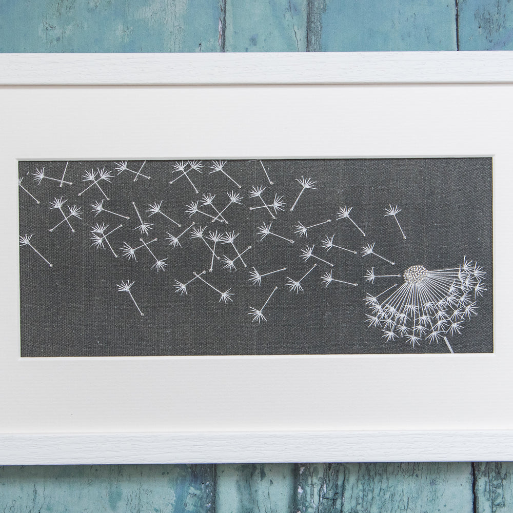 Framed finished embroidery of a dandelion clock caught in the wind on a charcoal cotton/linen background.