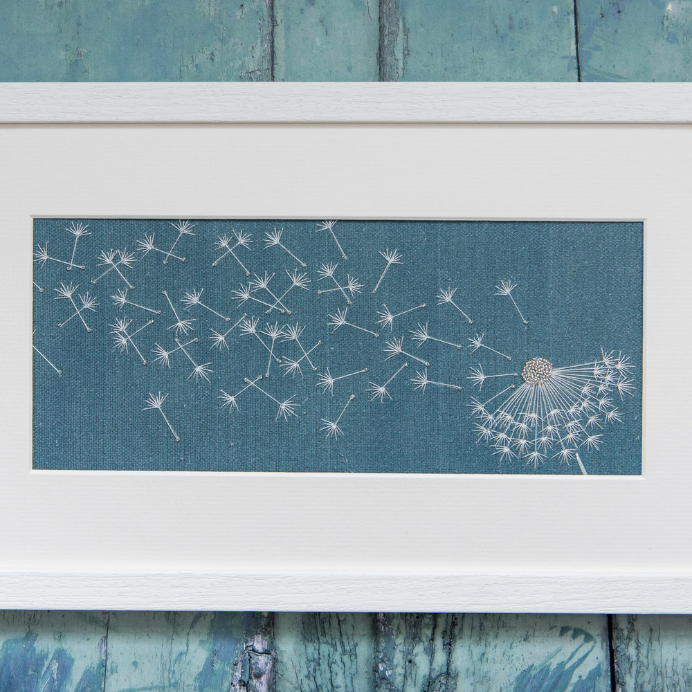 A petrol blue background with a embroidered dandelion clock blown by the wind. Framed in a white frame.