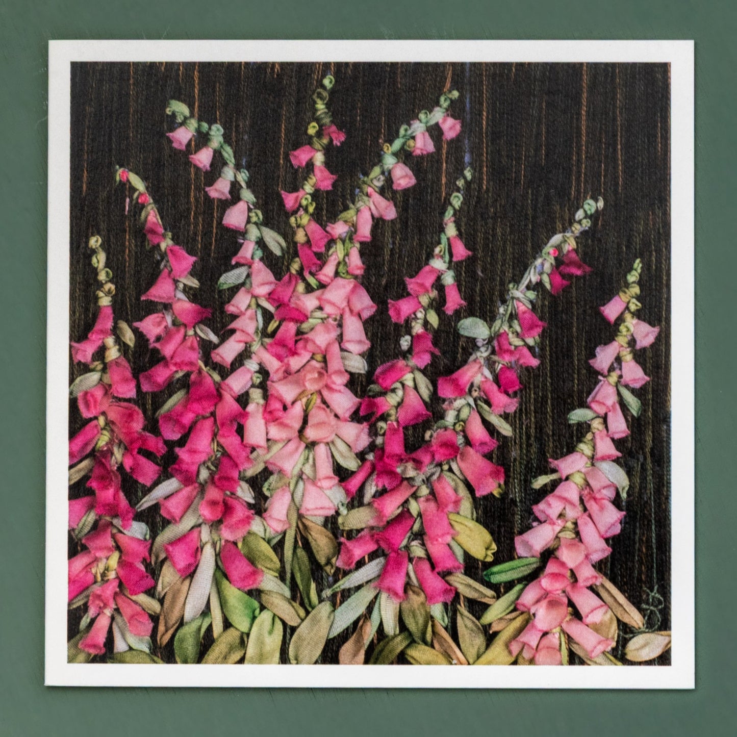 A card of ribbon embroidered pink foxgloves and their foliage. the Background is dark and embroidered using straight stitches in dark shades of brown with a few highlights.