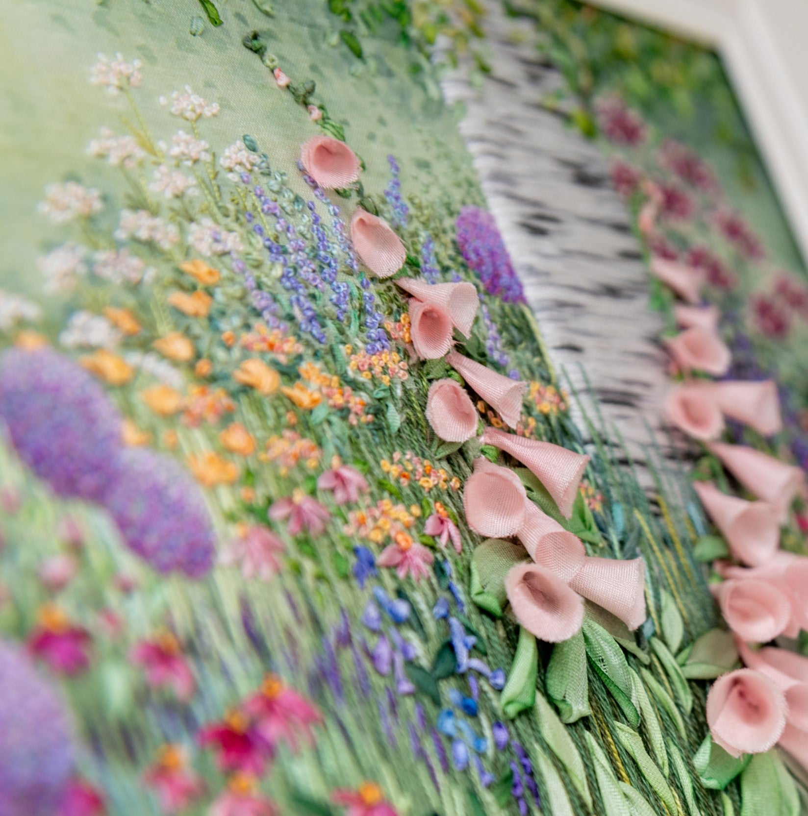 A close up of the hand embroidery titled mid summer border by Jo Butcher. The embroidery has a silver birch trunk rising from a summer flower border full of pale pink foxgloves, alliums and a range of pretty flowers.