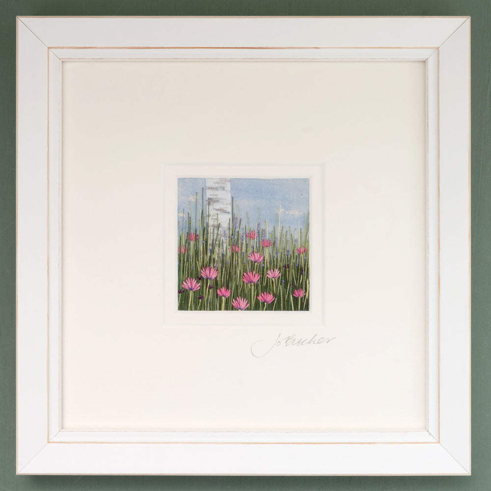 A white framed embroidery of a silver birch trunk rising above pretty pink flowers. Smaller purple and blue flowers nestle in the grasses.