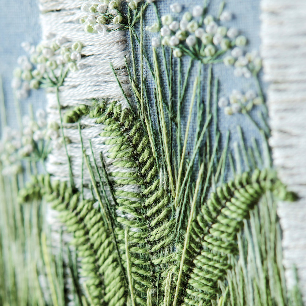 close up view of hand embroidered artwork of the bottom of a pair of silver birch trunks with cow parsley, ferns and grass