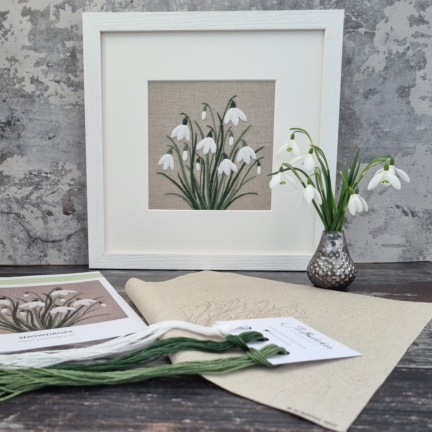 a white framed hand embroidered artwork of a clump of snowdrops. The art work is displayed on a shelf with a small vase of real snowdrops and the threads, cottons and background that comes with the kit