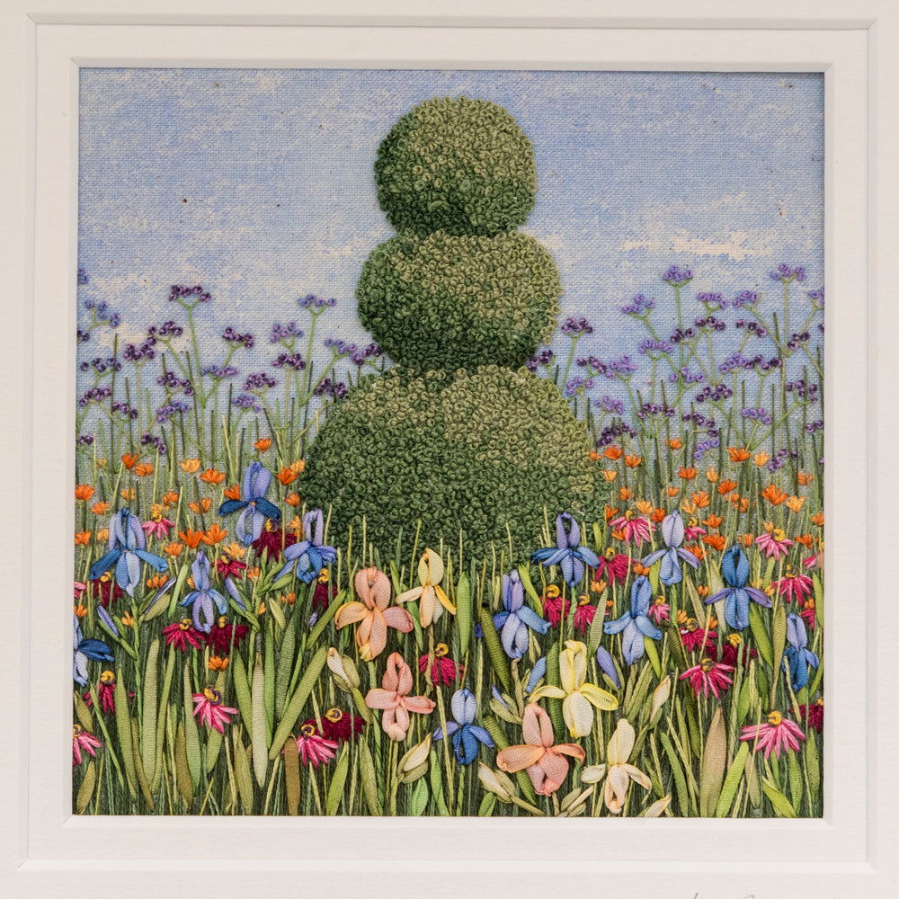 hand embroidery of a garden border with a topiary feature of three decreasing size balls on top of each other. The foreground shows Irises and foliage and verbena bonairensis gives some background height
