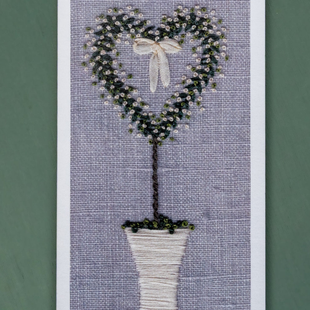 a hand embroidered white pot with a heart shaped tree with white roses and a ribbon bow hanging in the centre.