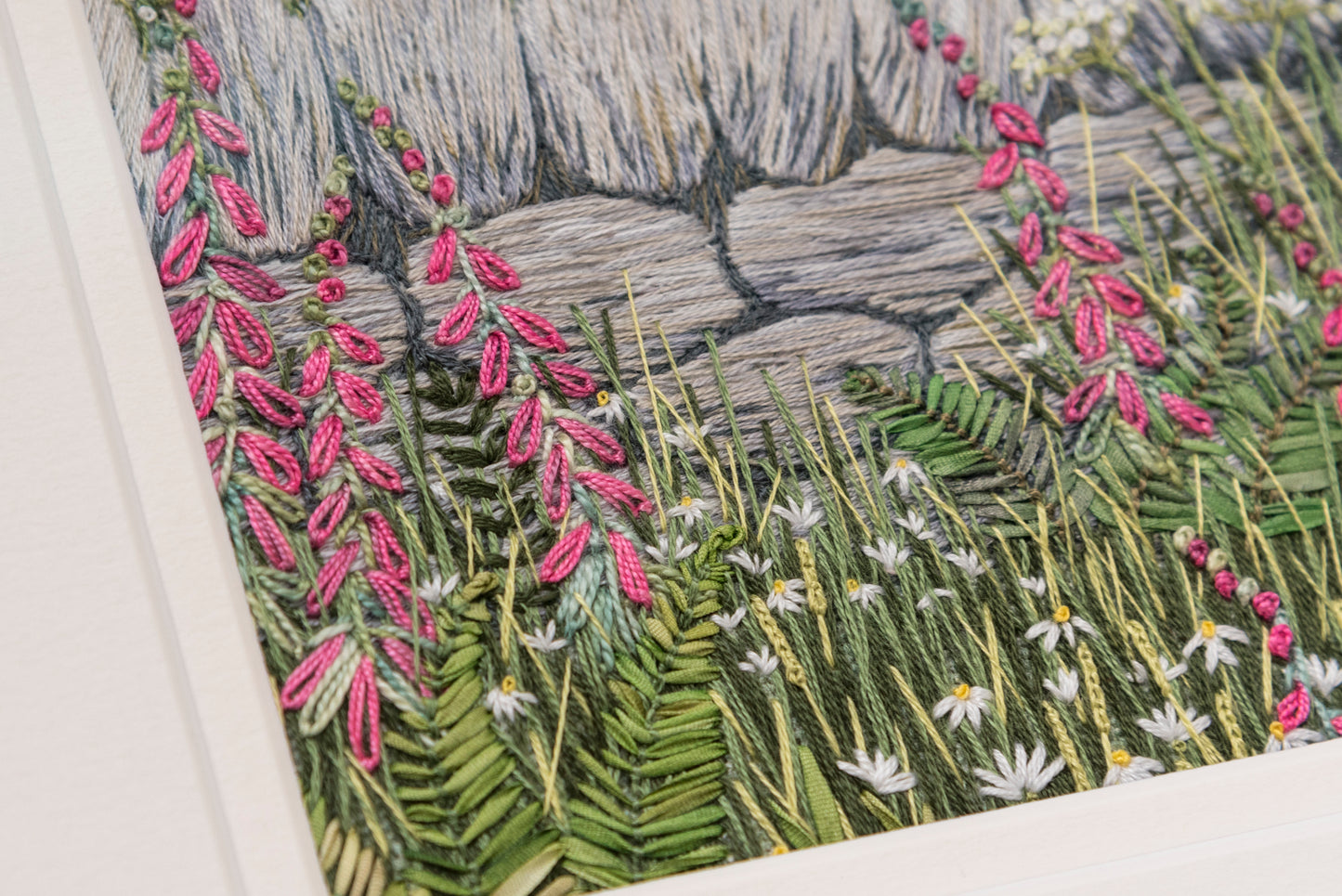 an angled close up of a hand embroidery artwork of a dry stone wall with pink foxgloves, ferns, daisy and grasses in front